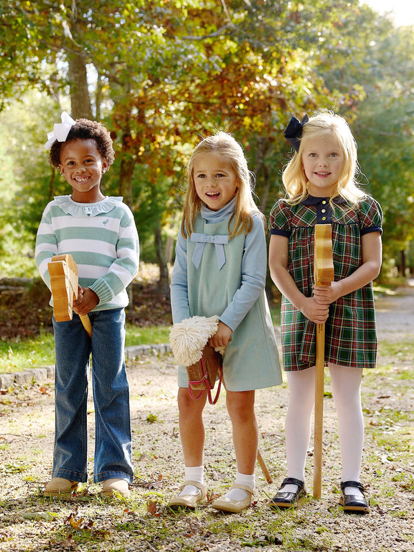 Three young girls stand outdoors on a path, smiling and holding hobby horse toys. They each wear different outfits: jeans with a sweater, a light blue dress, and The Beaufort Bonnet Company’s Dale Dress in Pinehill Plaid with Nantucket Navy—perfect for a girls’ fall dress look. Colorful autumn trees form the backdrop.
