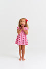 A young girl with curly hair and a red bow stands barefoot, smiling with her hands clasped, wearing The Beaufort Bonnet Company’s Penny’s Play Dress - Ladybug Line Up—a pink dress with a ladybug pattern—from the Ladybug Line Up collection, against a plain white background.