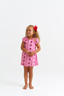 A young child with curly hair and a red bow smiles, standing barefoot in The Beaufort Bonnet Company’s Penny’s Play Dress - Ladybug Line Up, a light pink dress with red and black floral patterns, against a plain white background.