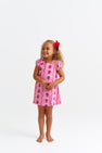 A young child with curly hair and a red bow smiles, standing barefoot in The Beaufort Bonnet Company’s Penny’s Play Dress - Ladybug Line Up, a light pink dress with red and black floral patterns, against a plain white background.