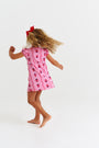 A young child with curly hair, barefoot, twirls against a plain white background wearing The Beaufort Bonnet Company’s Penny’s Play Dress - Ladybug Line Up, featuring a pink and red ladybug pattern with a red bow.