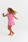 A young child with curly hair, barefoot, twirls against a plain white background wearing The Beaufort Bonnet Company’s Penny’s Play Dress - Ladybug Line Up, featuring a pink and red ladybug pattern with a red bow.