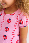 A smiling child with curly hair wears The Beaufort Bonnet Company’s Penny’s Play Dress - Ladybug Line Up, crafted from soft Pima Cotton and adorned with a charming print of red and navy ladybugs alongside small red flowers. Only the lower half of the face and upper torso are visible.