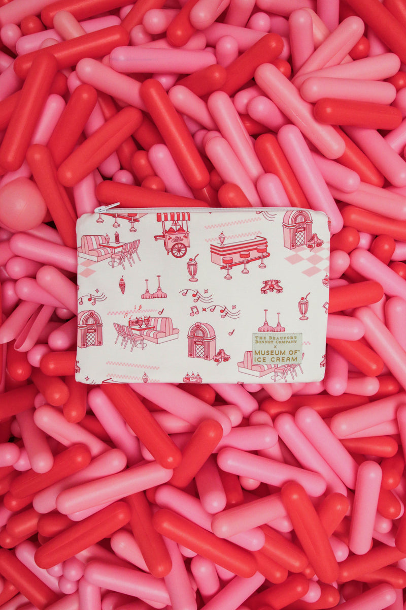 A charming retro print Dutchess Ditty Bag – Diner Days by The Beaufort Bonnet Company, featuring playful red illustrations on a white background, sits atop a pile of pink and red capsule-shaped objects—making it the perfect catchall for kids.