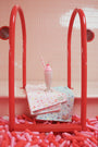 A pink and white milkshake with a cherry and straw sits atop pastel towels, including The Beaufort Bonnet Company’s Silent Night Throw in the Ice Cream Daydream (Blueberry Blue) print, draped over a red pool ladder. The scene is surrounded by pink and red balloon-like objects in a tiled room.