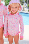 A young child wearing The Beaufort Bonnet Company’s Sarasota Surf Suit (UPF 35+) in Mableton Mini Floral (Pink) with Romany Rose and a matching sunhat stands by the pool, smiling at the camera. The sparkling water and lush greenery in the background showcase this stylish girls’ swimwear.