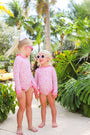 Two young children, both barefoot and smiling at each other, stand on a sunlit path surrounded by tropical plants and palm trees. They wear matching Sarasota Surf Suits (UPF 35+) in Mableton Mini Floral (Pink) with Romany Rose from The Beaufort Bonnet Company, paired with sunglasses.