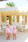 Two young children, both wearing The Beaufort Bonnet Company’s Sarasota Surf Suit (UPF 35+) in Mableton Mini Floral (Pink) with Romany Rose, stand together by white metal patio furniture under a yellow-and-white canopy on a sunny day.