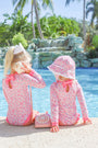 Two young girls in matching The Beaufort Bonnet Company Sarasota Surf Suits (UPF 35+) in Mableton Mini Floral (Pink) with Romany Rose sit by a pool. One holds a seashell to her ear while the other wears a sunhat. A pink vintage rotary phone rests between them, with palm trees and a waterfall in the background.