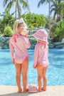 Two young children wearing matching Sarasota Surf Suits (UPF 35+) in Mableton Mini Floral (Pink) with Romany Rose by The Beaufort Bonnet Company stand by a pool. One holds a seashell to her ear while the other wears a sunhat. A pink toy phone lies on the ground between them, surrounded by palm trees and lush greenery in the background.