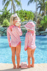 Two young children wearing matching Sarasota Surf Suits (UPF 35+) in Mableton Mini Floral (Pink) with Romany Rose by The Beaufort Bonnet Company stand by a pool. One holds a seashell to her ear while the other wears a sunhat. A pink toy phone lies on the ground between them, surrounded by palm trees and lush greenery in the background.