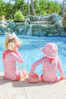 Two young girls in matching Sarasota Surf Suits (UPF 35+) in the Mableton Mini Floral (Pink) with Romany Rose print from The Beaufort Bonnet Company sit by a pool. One listens to a seashell, while the other touches a pink rotary phone. With a backdrop of cascading water and palm trees, the scene captures the playful charm of adorable girls swimwear styles.
