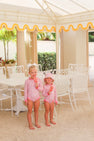 Two young girls, smiling and holding popsicles, stand by a white patio table and chairs under a yellow-trimmed canopy. They are dressed in matching Sarasota Surf Suits (UPF 35+) in Mableton Mini Floral (Pink) with Romany Rose by The Beaufort Bonnet Company, complete with coordinating hats.