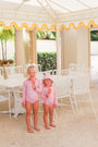 Two young children in matching The Beaufort Bonnet Company Sarasota Surf Suits (UPF 35+) in Mableton Mini Floral (Pink) with Romany Rose hold pink ice cream bars, smiling under a white and yellow striped canopy next to white patio chairs and a table on a stone patio.