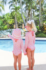 Two young girls in matching Sarasota Surf Suits (UPF 35+) in Mableton Mini Floral (Pink) with Romany Rose from The Beaufort Bonnet Company stand by the pool, holding hands. One points toward the water while the other, wearing a white bow, gazes ahead. Palm trees and lush greenery set the scene—an adorable summer moment highlighting stylish swimwear for girls.