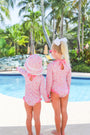 Two young children wearing matching Sarasota Surf Suits (UPF 35+) in Mableton Mini Floral (Pink) with Romany Rose by The Beaufort Bonnet Company stand by a pool, holding hands. One wears a sunhat, the other a white bow. Palm trees and lush greenery are in the background on a sunny day.