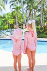 Two young children wearing matching Sarasota Surf Suits (UPF 35+) in Mableton Mini Floral (Pink) with Romany Rose by The Beaufort Bonnet Company stand by a pool, holding hands. One wears a sunhat, the other a white bow. Palm trees and lush greenery are in the background on a sunny day.