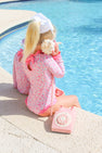 A young girl wearing The Beaufort Bonnet Company’s Sarasota Surf Suit (UPF 35+) in Mableton Mini Floral (Pink) with Romany Rose sits by a pool, holding a seashell to her ear, with a vintage pink rotary phone beside her.