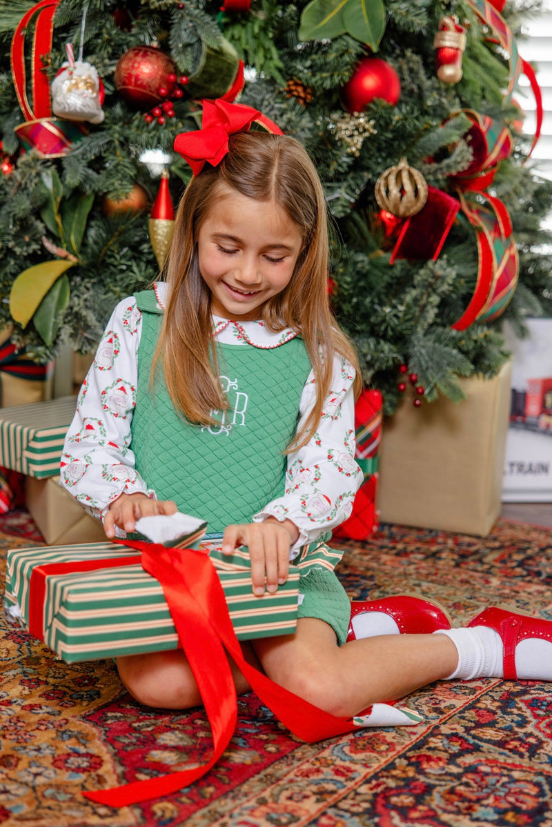 A young girl dressed in The Beaufort Bonnet Company’s Long Sleeve Maude’s Peter Pan Collar Shirt in North Pole Portrait with Richmond Red, with a festive red bow in her hair, opens a striped gift box in front of a decorated Christmas tree adorned with wrapped presents underneath.