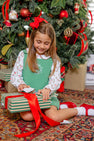 A young girl dressed in The Beaufort Bonnet Company’s Long Sleeve Maude’s Peter Pan Collar Shirt in North Pole Portrait with Richmond Red, with a festive red bow in her hair, opens a striped gift box in front of a decorated Christmas tree adorned with wrapped presents underneath.