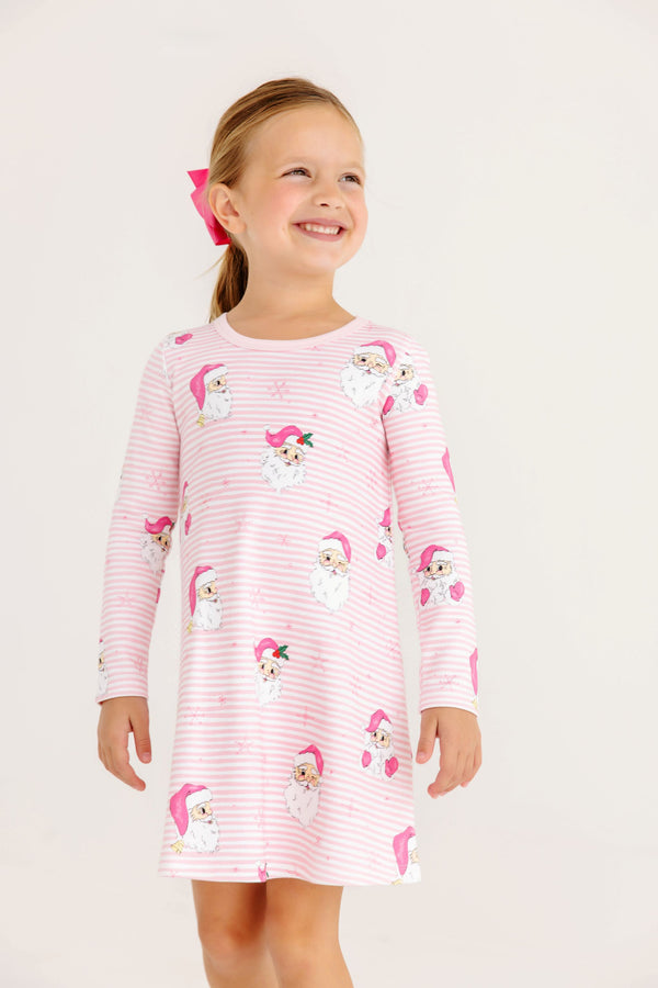 A smiling girl wears a Long Sleeve Polly Play Dress - Dear Santa (Palm Beach Pink Stripe) with Palm Beach Pink, featuring pink and white stripes and a pattern of Santa Claus faces.