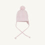The Parrish Pom Pom Hat - Palm Beach Pink by The Beaufort Bonnet Company is a light pink cotton knit hat showcasing a classic cable-knit pattern, playful pom-pom on top, ear flaps on each side, and two braided tassels, all presented against a plain white background.