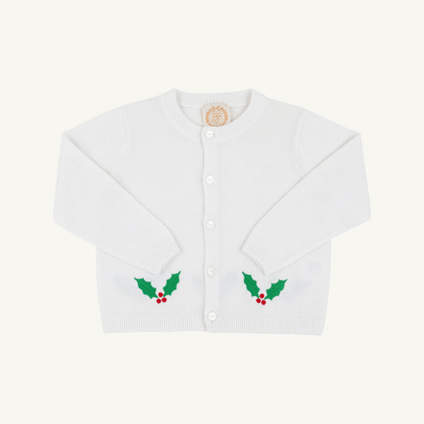 The Beaufort Bonnet Company’s Cambridge Cardigan in Worth Avenue White features long sleeves, front buttons, and festive green holly leaf with red berry designs near the bottom on each side. This cheerful baby sweater stands out against its plain light background, making it a perfect choice for the holiday season.