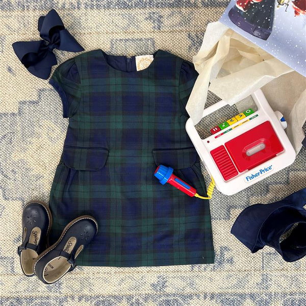A The Beaufort Bonnet Company Betts Bow Dress - Berwick Black Watch with Nantucket Navy Velveteen Bows is arranged alongside a toy microphone and a Fisher-Price cash register on a blue-patterned rug, with tissue paper and a box placed in the corner.