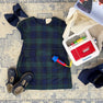 A The Beaufort Bonnet Company Betts Bow Dress - Berwick Black Watch with Nantucket Navy Velveteen Bows is arranged alongside a toy microphone and a Fisher-Price cash register on a blue-patterned rug, with tissue paper and a box placed in the corner.