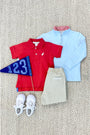 Children’s outfit laid out on a textured carpet: The Beaufort Bonnet Company Hayword Half-Zip in Buckhead Blue with Richmond Red Stripe, beige pants, white sneakers, and a blue 123 flag.