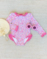A Sarasota Surf Suit (UPF 35+) in Mableton Mini Floral (Pink) with Romany Rose by The Beaufort Bonnet Company, pink round sunglasses, and a woven straw bag are arranged on a textured beige rug.