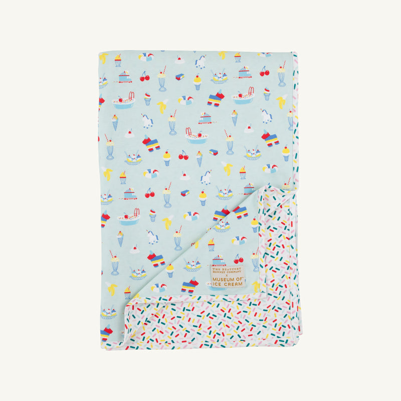 A folded Silent Night Throw - Ice Cream Daydream (Blueberry Blue) by The Beaufort Bonnet Company, featuring playful colorful ice cream and cherry illustrations on a light blue background, partially revealing a sprinkle-patterned reverse side. The Museum of Ice Cream label is visible on the front.