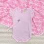 A pale pink baby bodysuit, a Parker Paci Clip - Pink Greenbrier Gingham with Palm Beach Pink, and a pink blanket with a white bow pattern.