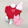 Flat lay of a child’s outfit: red sweater with a white bow design, white mock neck shirt, The Beaufort Bonnet Company’s Collins Cable Knit Hat in Palm Beach Pink, light pink leggings, and white lace-up shoes on a textured cream rug.
