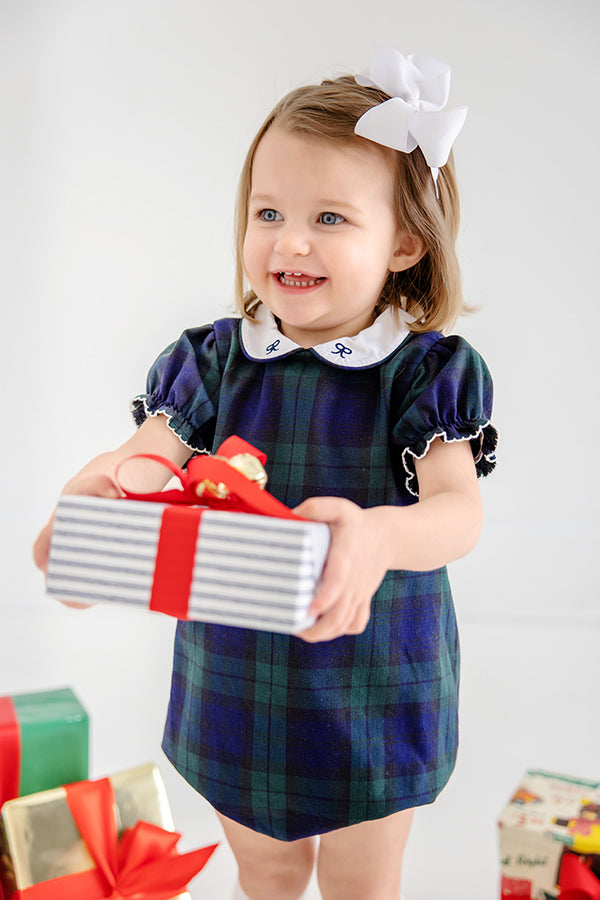 A smiling toddler in The Beaufort Bonnet Company’s Bedford Bubble - Berwick Black Watch with Worth Avenue White & Nantucket Navy Bows dress, with a white bow in her hair, holds a gift wrapped in striped paper with a red ribbon. More presents are visible at her feet.
