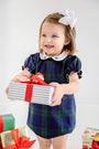 A smiling toddler in The Beaufort Bonnet Company’s Bedford Bubble - Berwick Black Watch with Worth Avenue White & Nantucket Navy Bows dress, with a white bow in her hair, holds a gift wrapped in striped paper with a red ribbon. More presents are visible at her feet.