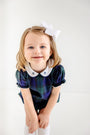 A young girl with light brown hair, wearing The Beaufort Bonnet Company’s Bedford Bubble in Berwick Black Watch with Worth Avenue White & Nantucket Navy Bows, smiles and leans slightly forward against a white background.