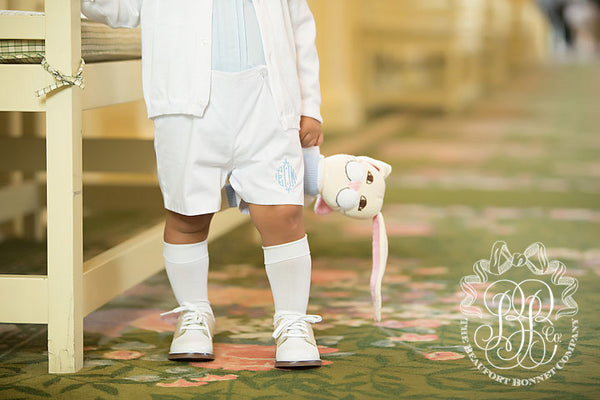 A young child stands indoors, holding a plush bunny toy by its ear. The child is dressed in classic style, wearing white shorts, dress shoes, and Jefferies High Class Knee Socks by The Beaufort Bonnet Company. The child’s face is not visible. This timeless look is perfect as a T.B.B.C. outfit for girls or boys.