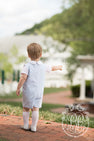 A young child in a light blue outfit from The Beaufort Bonnet Company and Jefferies High Class Knee Socks in White stands on a brick path, pointing into the distance with their back to the camera. Trees and a white building are visible in the background. This charming look from The Beaufort Bonnet Company is perfect for girls or boys.