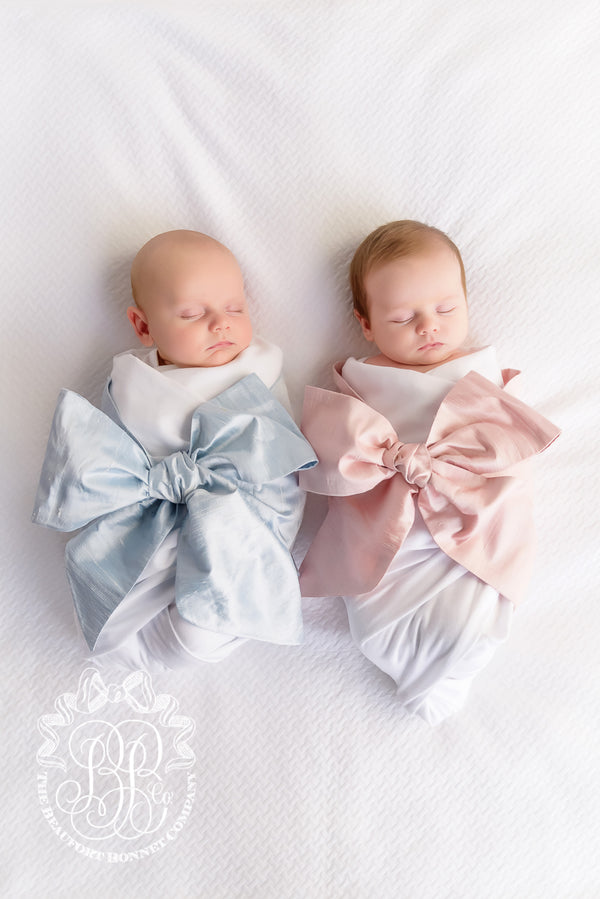 Two newborns in a Bow Swaddle ® - Southern Blush Silk, each wrapped in a white blanket with a large, decorative bow in light blue or soft pink, perfect for baby keepsake photos.