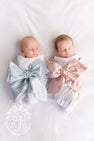 Two newborns in a Bow Swaddle ® - Southern Blush Silk, each wrapped in a white blanket with a large, decorative bow in light blue or soft pink, perfect for baby keepsake photos.