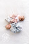 Two infants lie head-to-toe in Bow Swaddle ® - Southern Blush Silk, both swaddled in white fabric with large, oversized bows in light pink and dusty blue to create a perfect newborn picture.