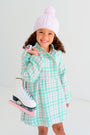 A smiling young girl wears The Beaufort Bonnet Company’s Collins Cable Knit Hat in Palm Beach Pink and a green plaid dress, holding white ice skates with pink blade guards over her shoulder, standing against a plain white background.