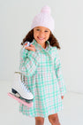 A smiling young girl wears The Beaufort Bonnet Company’s Collins Cable Knit Hat in Palm Beach Pink and a green plaid dress, holding white ice skates with pink blade guards over her shoulder, standing against a plain white background.