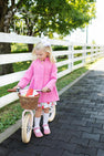 A young girl wearing The Beaufort Bonnet Company’s Penelope Peacoat in Hamptons Hot Pink with Richmond Red and a white bow stands next to a small bicycle with a basket, on a paved path by a white fence and green trees, looking down.