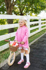 A young blonde girl wearing The Beaufort Bonnet Company’s Penelope Peacoat in Hamptons Hot Pink with Richmond Red, a white patterned dress, and white knee-high socks stands next to a pink balance bike with a wicker basket filled with wrapped gifts. She has a white bow in her hair and is standing by a white fence.