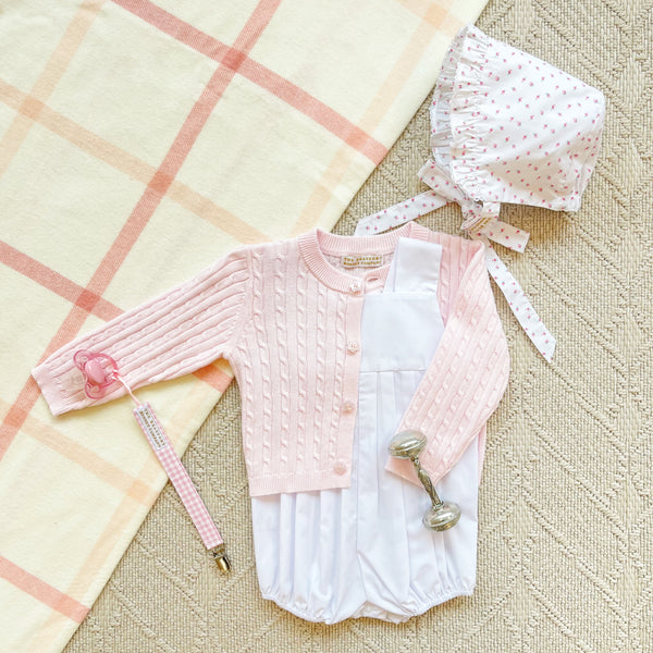 A pink cable-knit cardigan, white bubble romper, matching floral bonnet, silver rattle, and a Parker Paci Clip - Pink Greenbrier Gingham with Palm Beach Pink pacifier, all arranged on a plaid blanket.