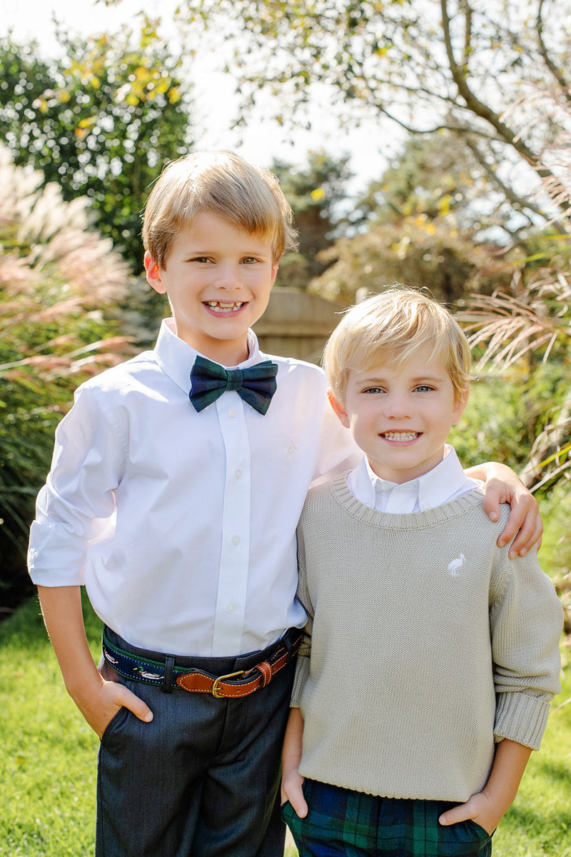 Two young boys stand close together outdoors, smiling at the camera. One wears The Beaufort Bonnet Company’s Baylor Bowtie - Berwick Black Watch with his white shirt, while the other sports a beige sweater. Sunlight and greenery are visible in the background.