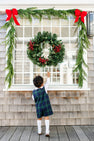 Dressed in the Jack Keene Jon Jon - Berwick Black Watch by The Beaufort Bonnet Company, a young child from the little gents wardrobe points up at a decorated window adorned with a Christmas wreath and garland, both tied with red bows for a festive, bright scene.