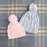 Two knitted beanies with pom-poms, including The Beaufort Bonnet Company’s Collins Cable Knit Hat in Palm Beach Pink and a light blue one, are placed on a blue and white plaid fabric background.